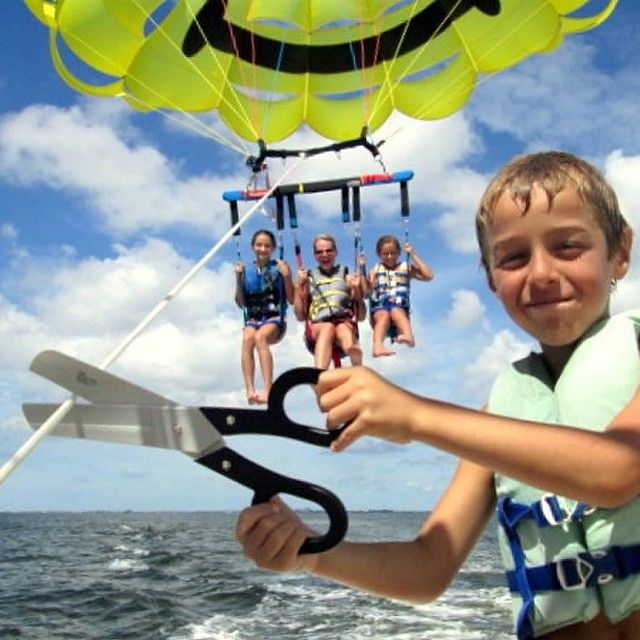 Boy on a boat holding scissors-like handle while three people parasail overhead above the ocean.