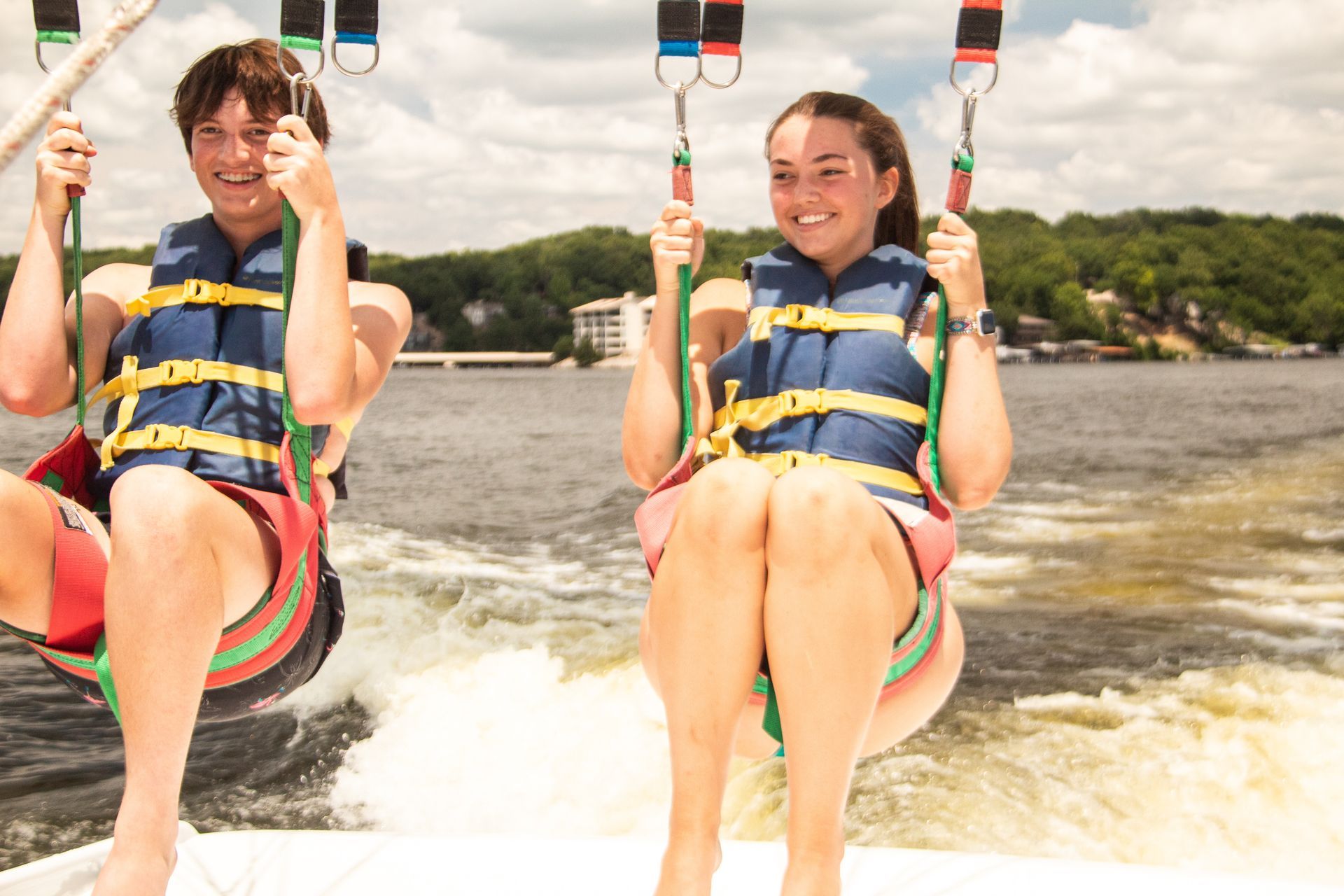 Two people in life jackets ride a towable tube behind a boat, smiling on the water.