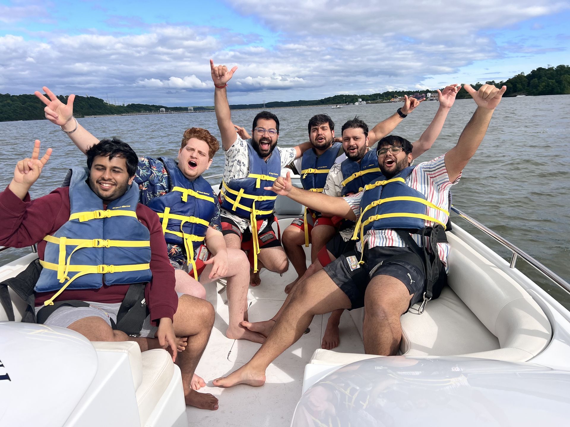 Six friends in life vests smiling and posing on a boat on a lake with arms raised