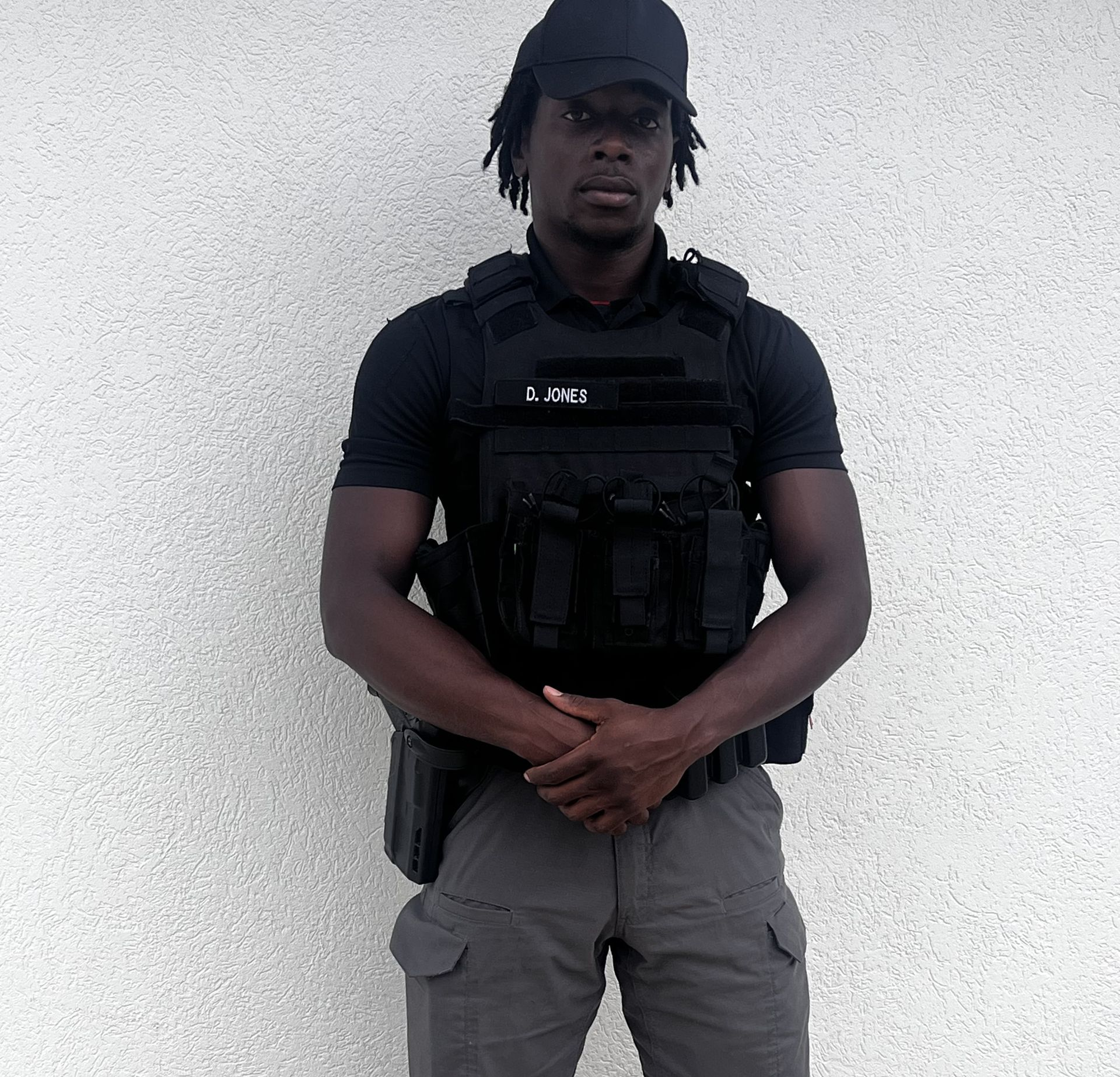 Man in black tactical gear stands against a white wall.