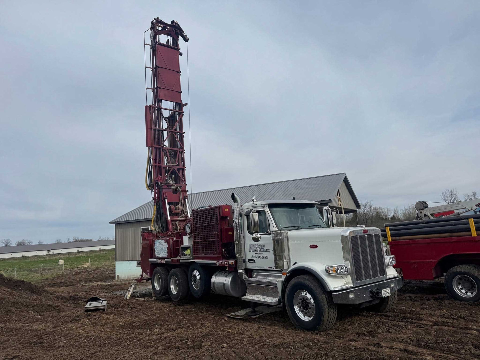 White truck with a tall red drilling rig parked on a dirt lot beside a barn under an overcast sky