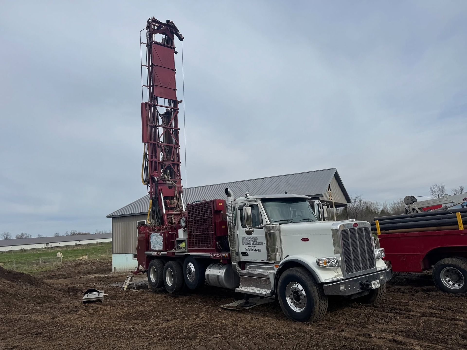 White semi-truck with a tall red drilling rig parked beside a building in a rural field.