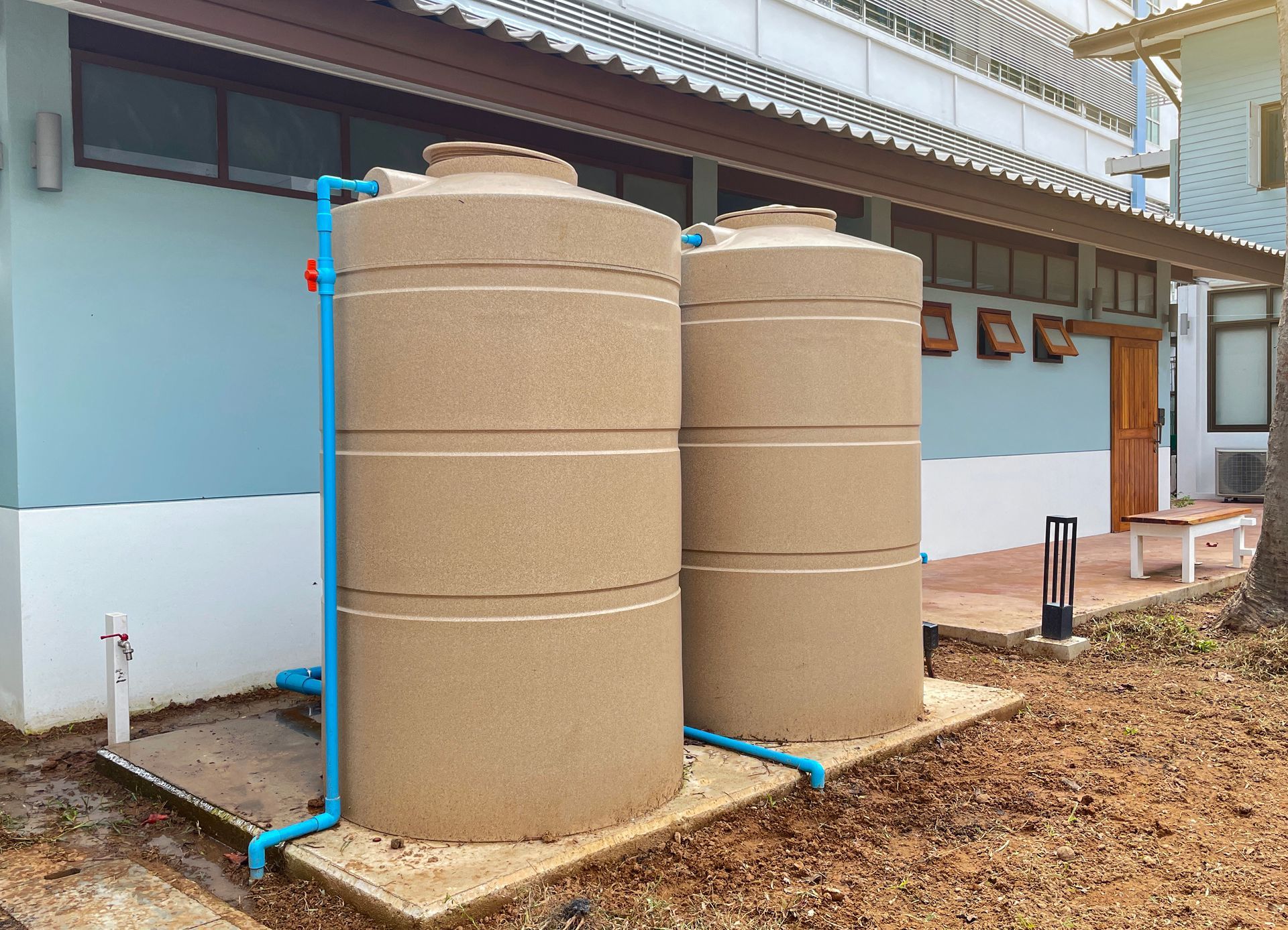 Two beige cylindrical water tanks beside a light blue building, connected by blue pipes.