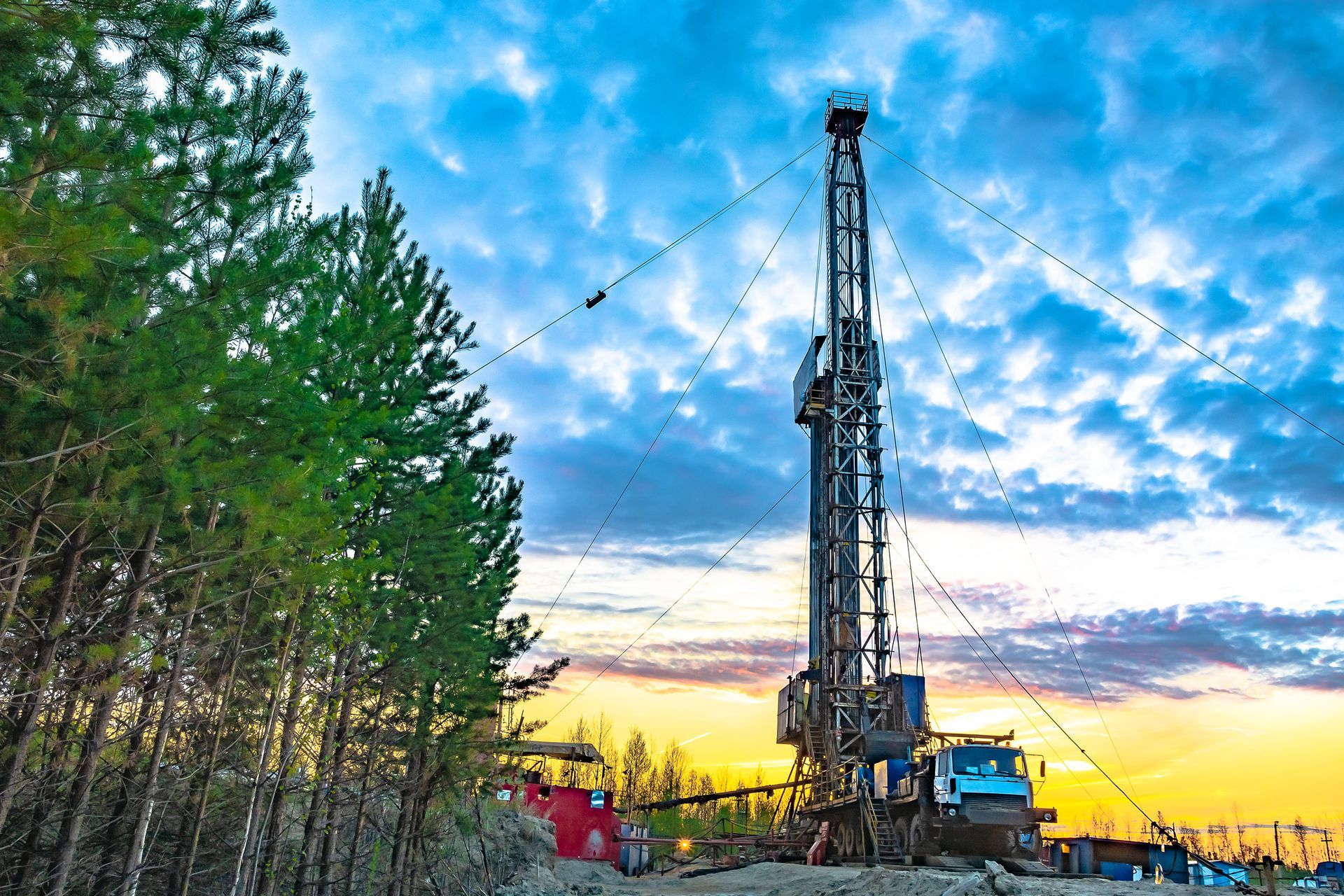 Oil drilling rig at sunset beside pine trees under a blue, clouded sky
