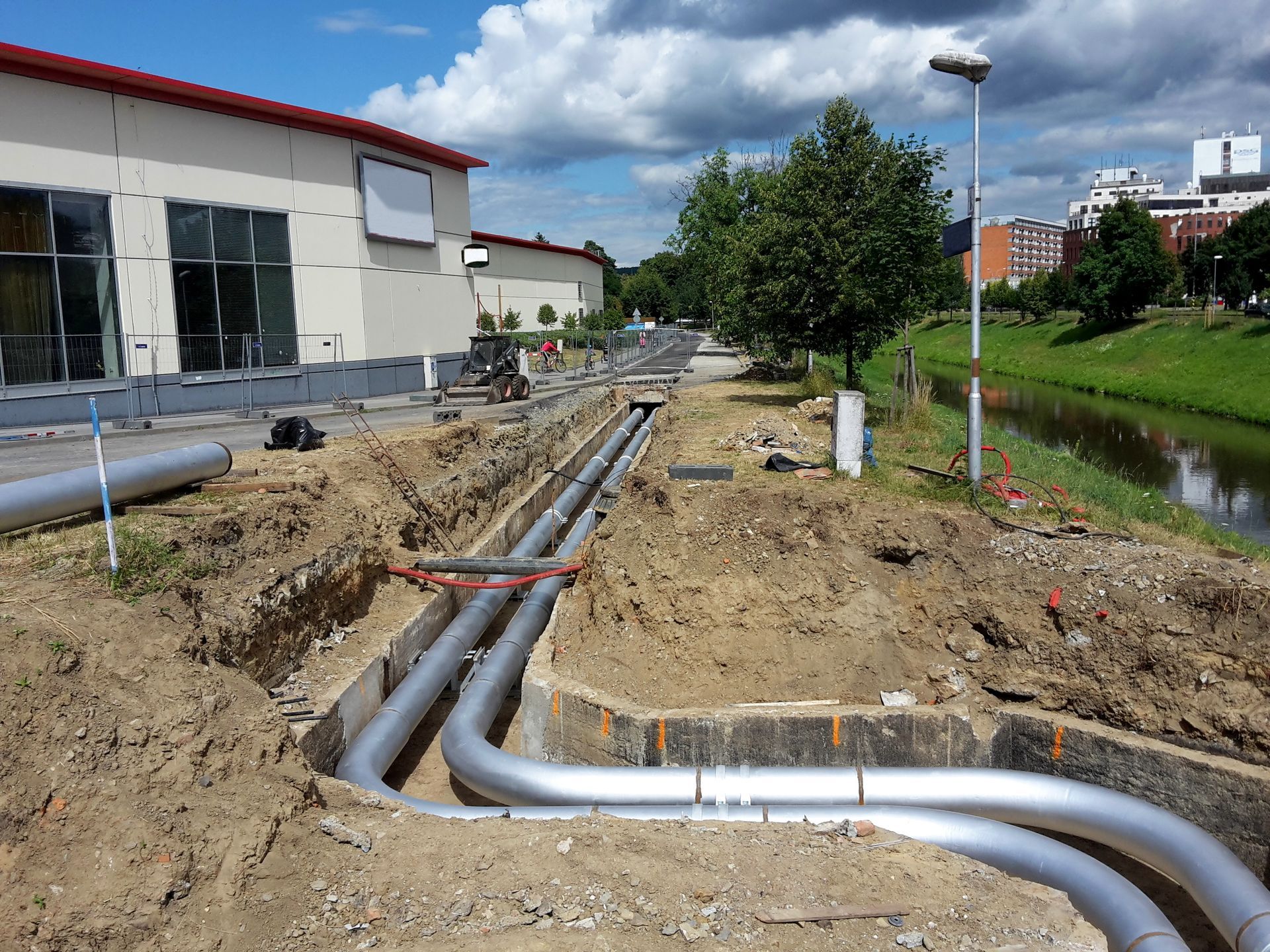 Construction site with large silver utility pipes laid in a trench beside a building and canal.