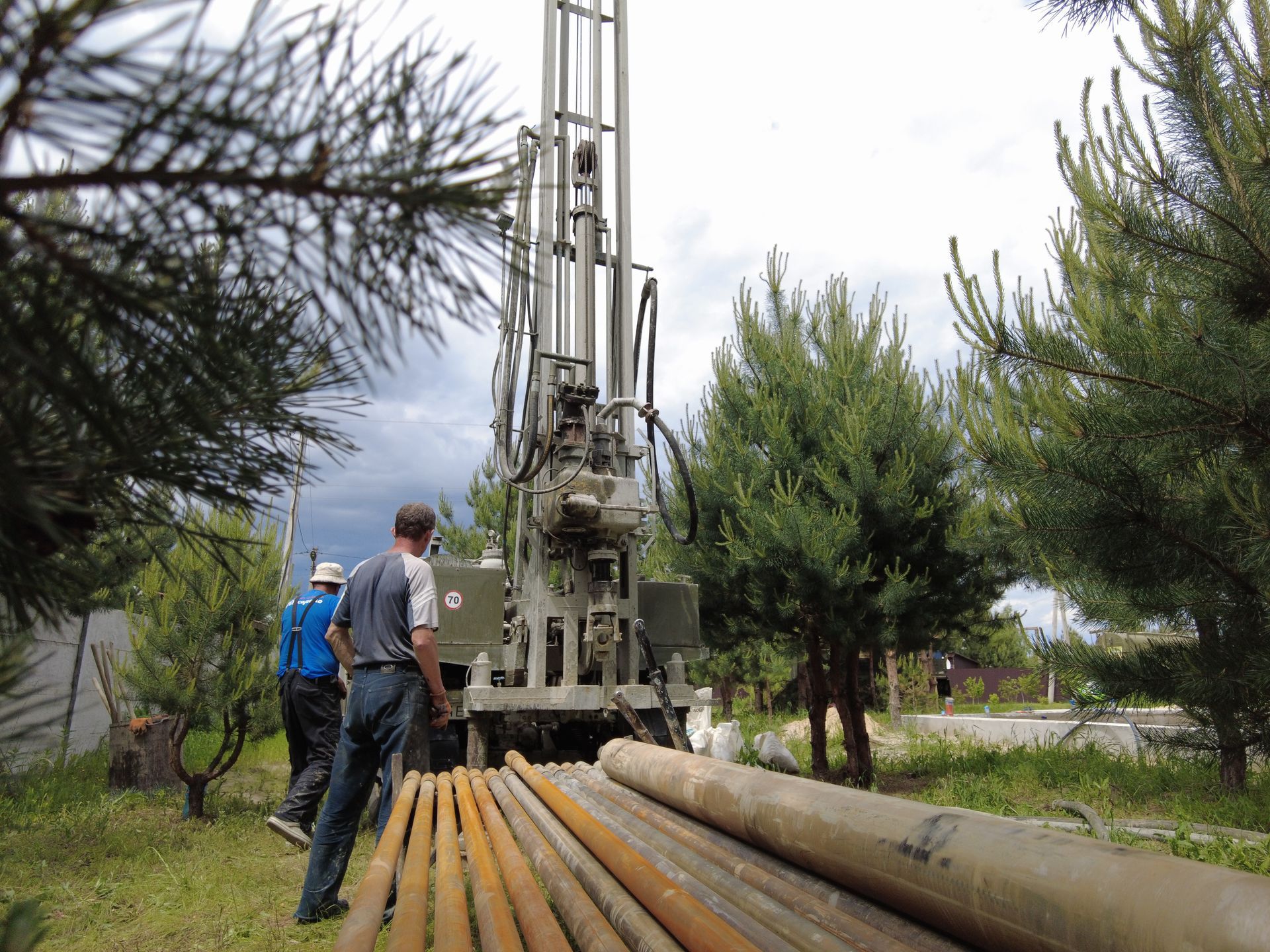 Workers beside a drilling rig and stacked pipes in a wooded outdoor area