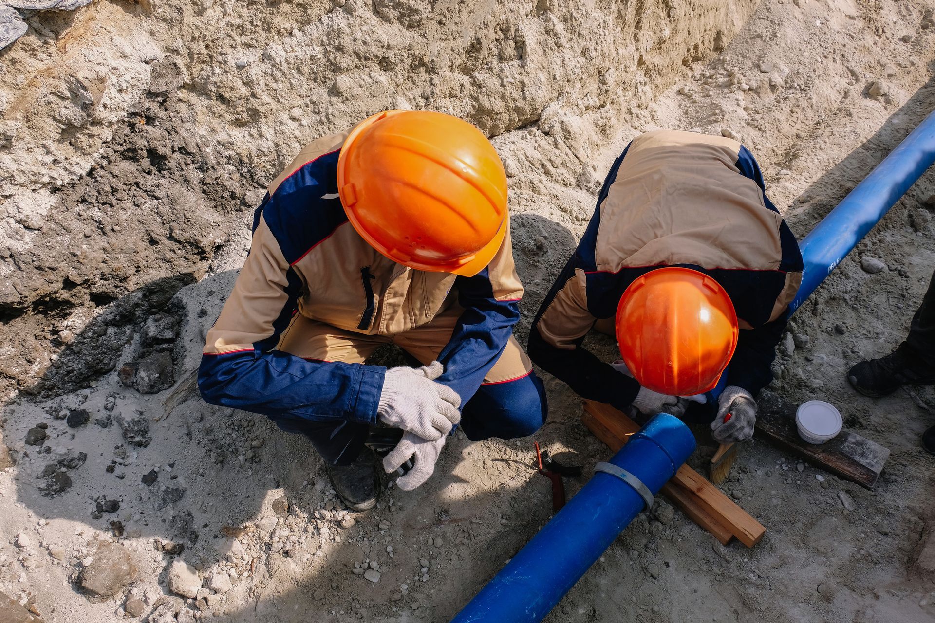 Two workers in orange hard hats repair a blue pipe in a dirt trench.