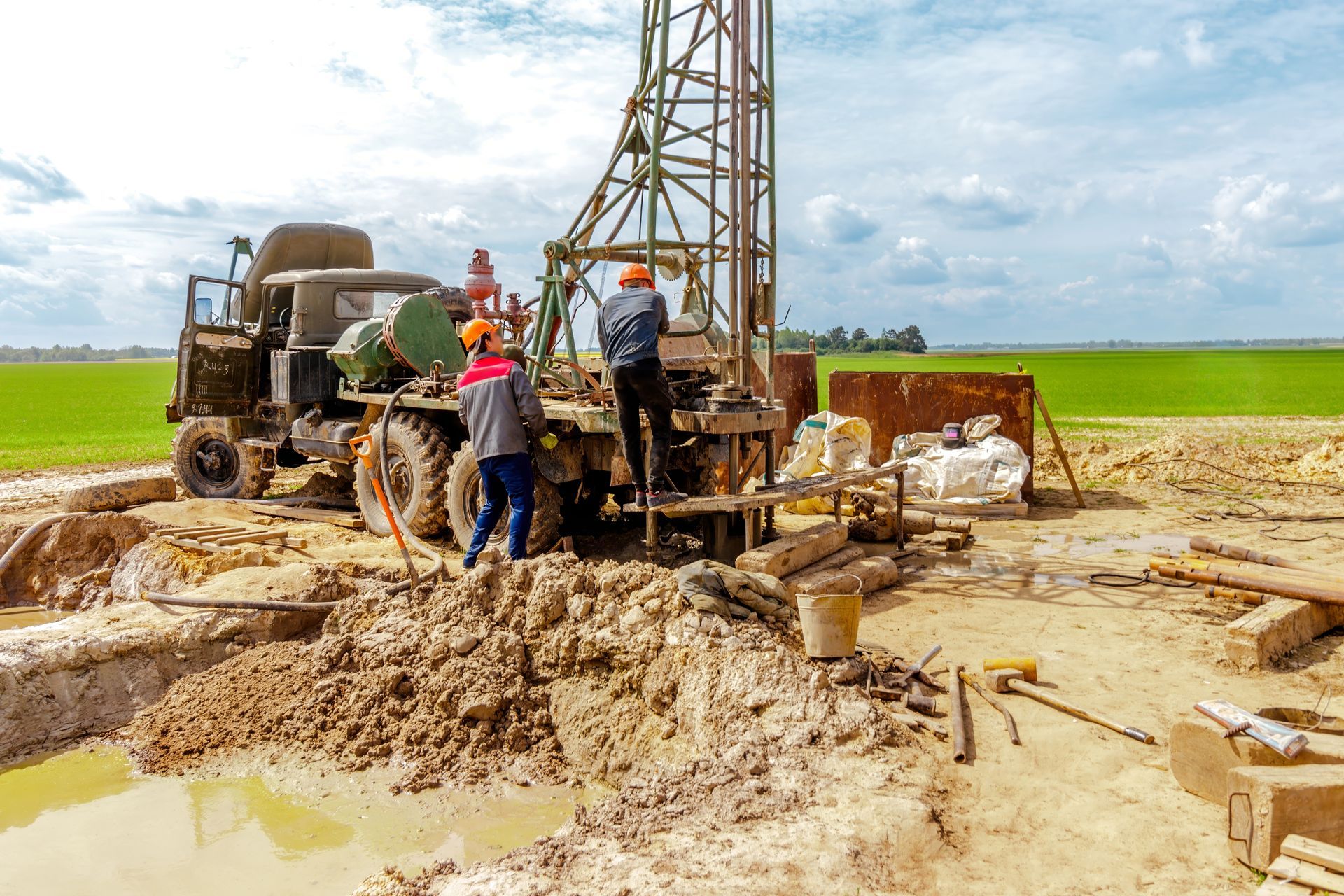 Workers operating drilling machinery at a muddy construction site under a cloudy sky