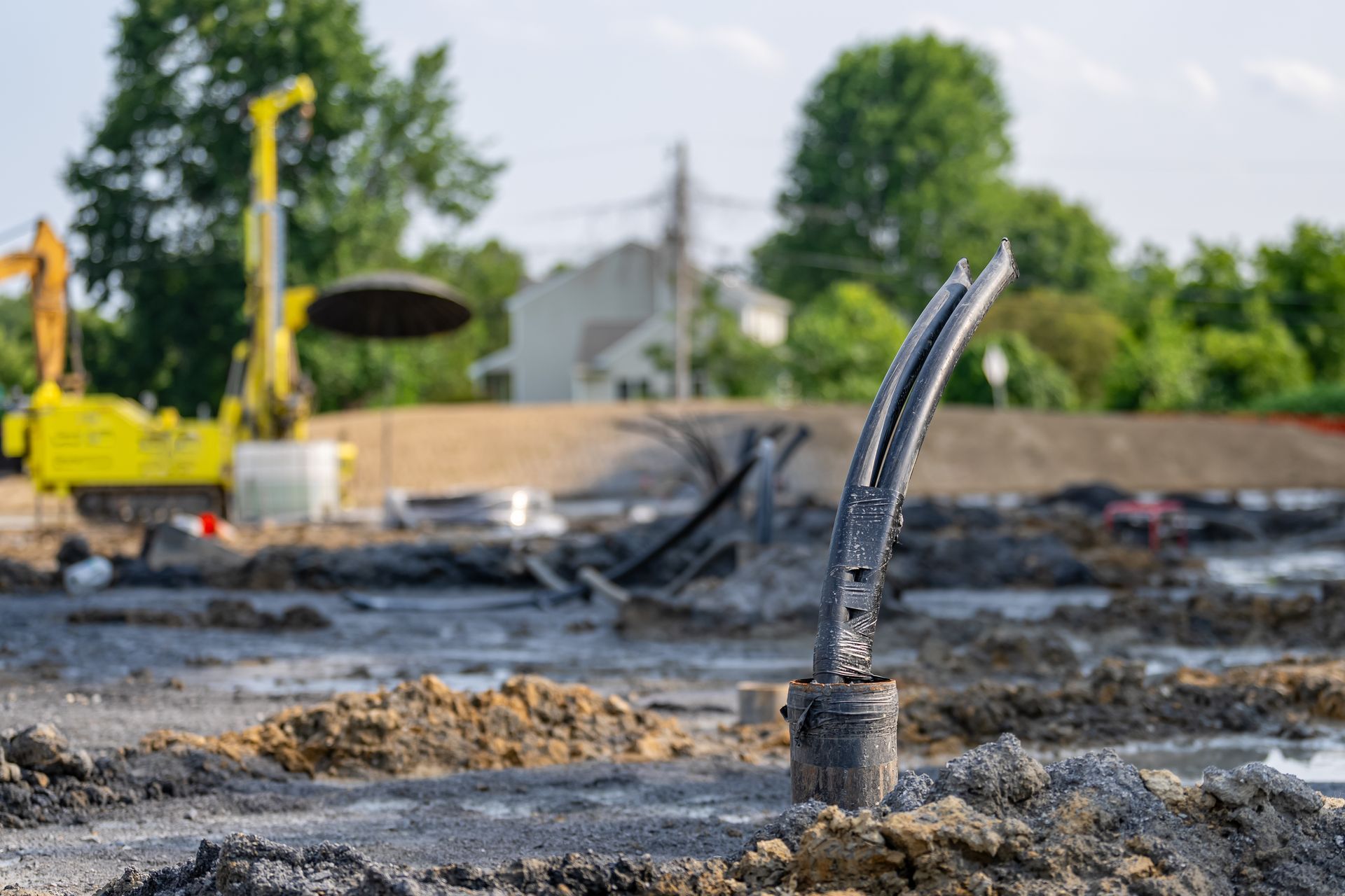 Construction site with exposed pipes and muddy ground, yellow excavator in the background.