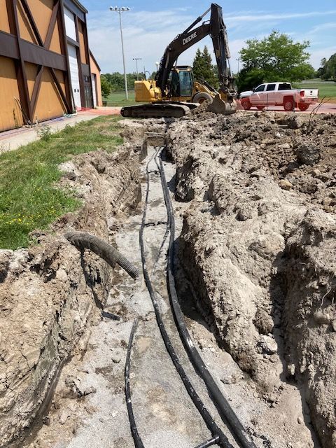 Construction trench beside building with exposed black cables and excavator in the background