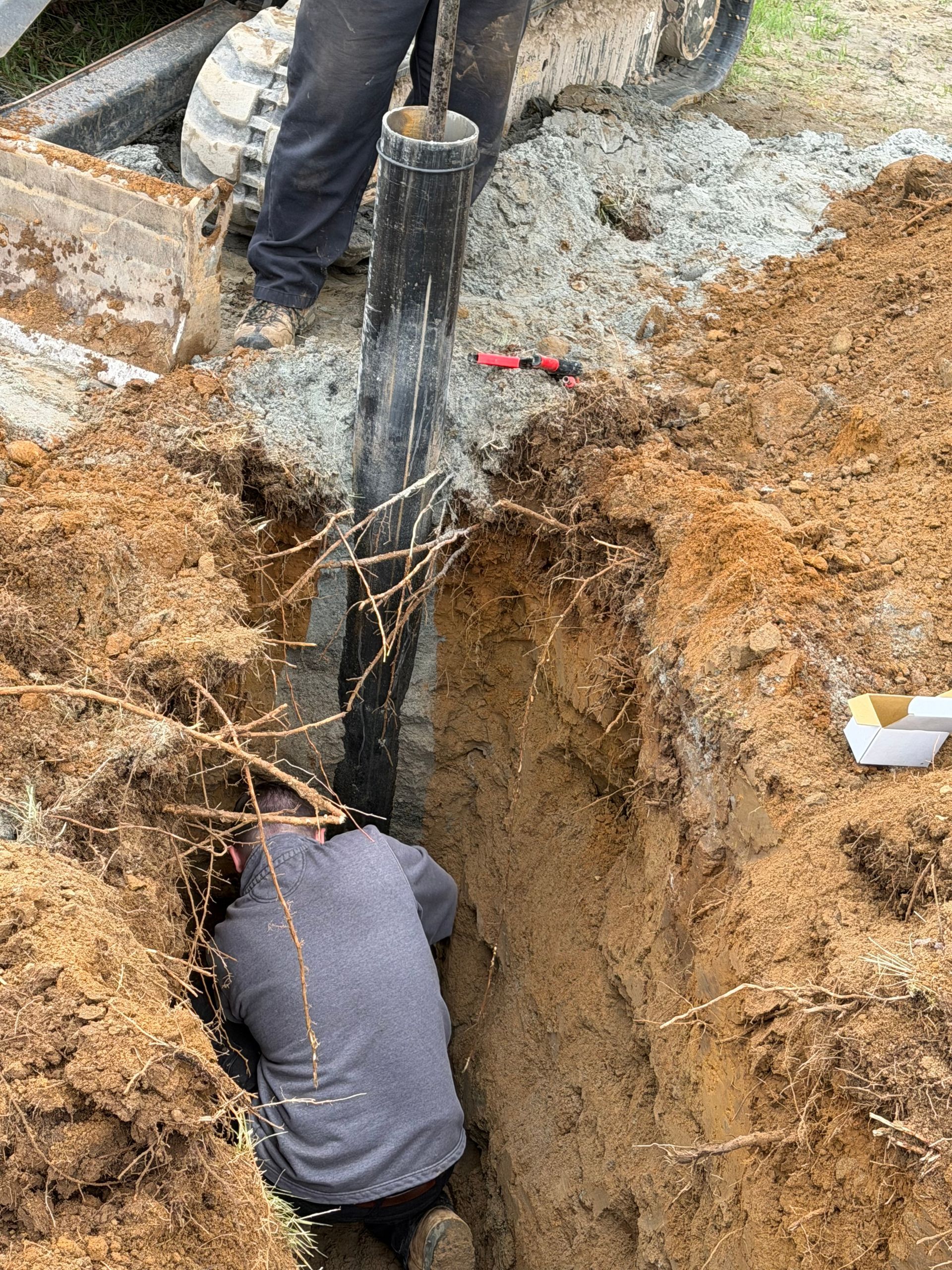 Worker in a deep trench inspecting a cable or pipe beside a utility pole and excavated dirt.
