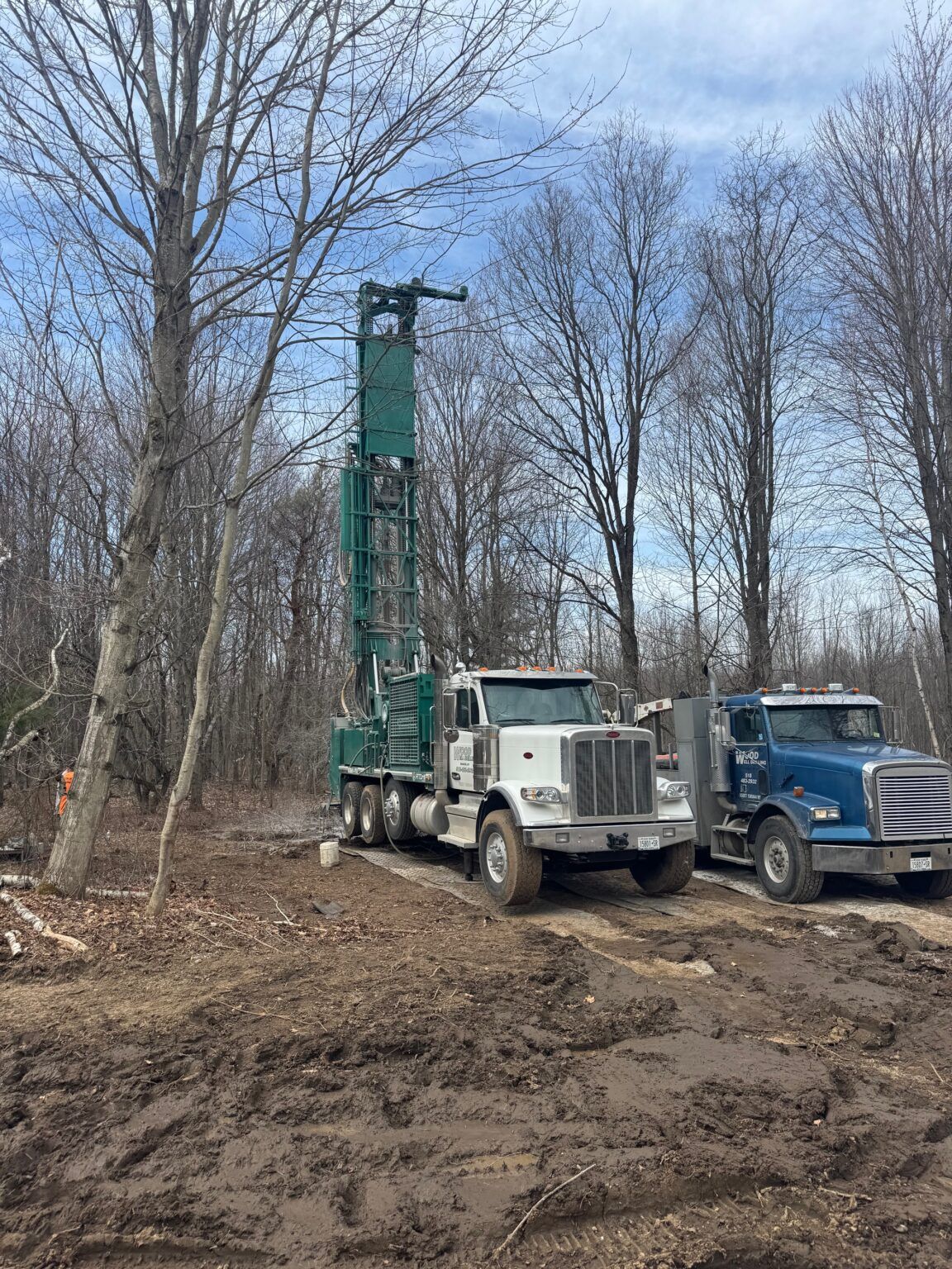 Two drilling trucks in a muddy wooded clearing under bare trees and a blue sky