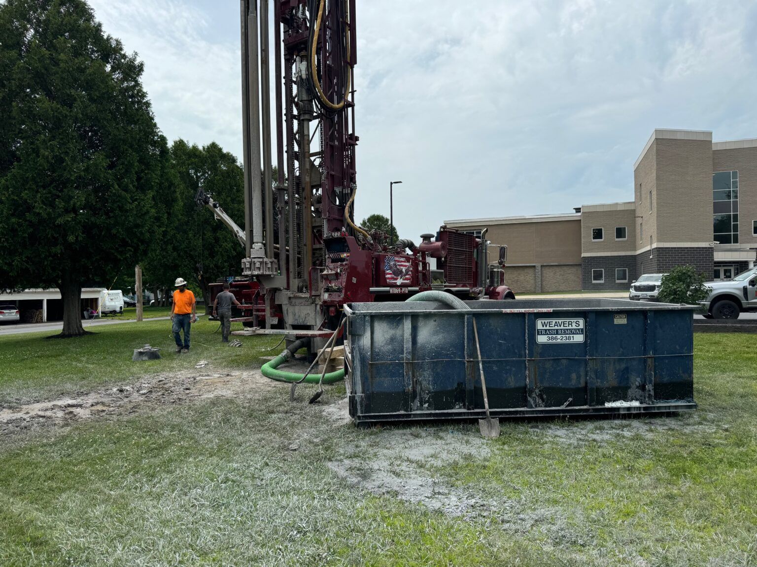 Workers beside drilling rig and blue dumpster at a construction site near a building.