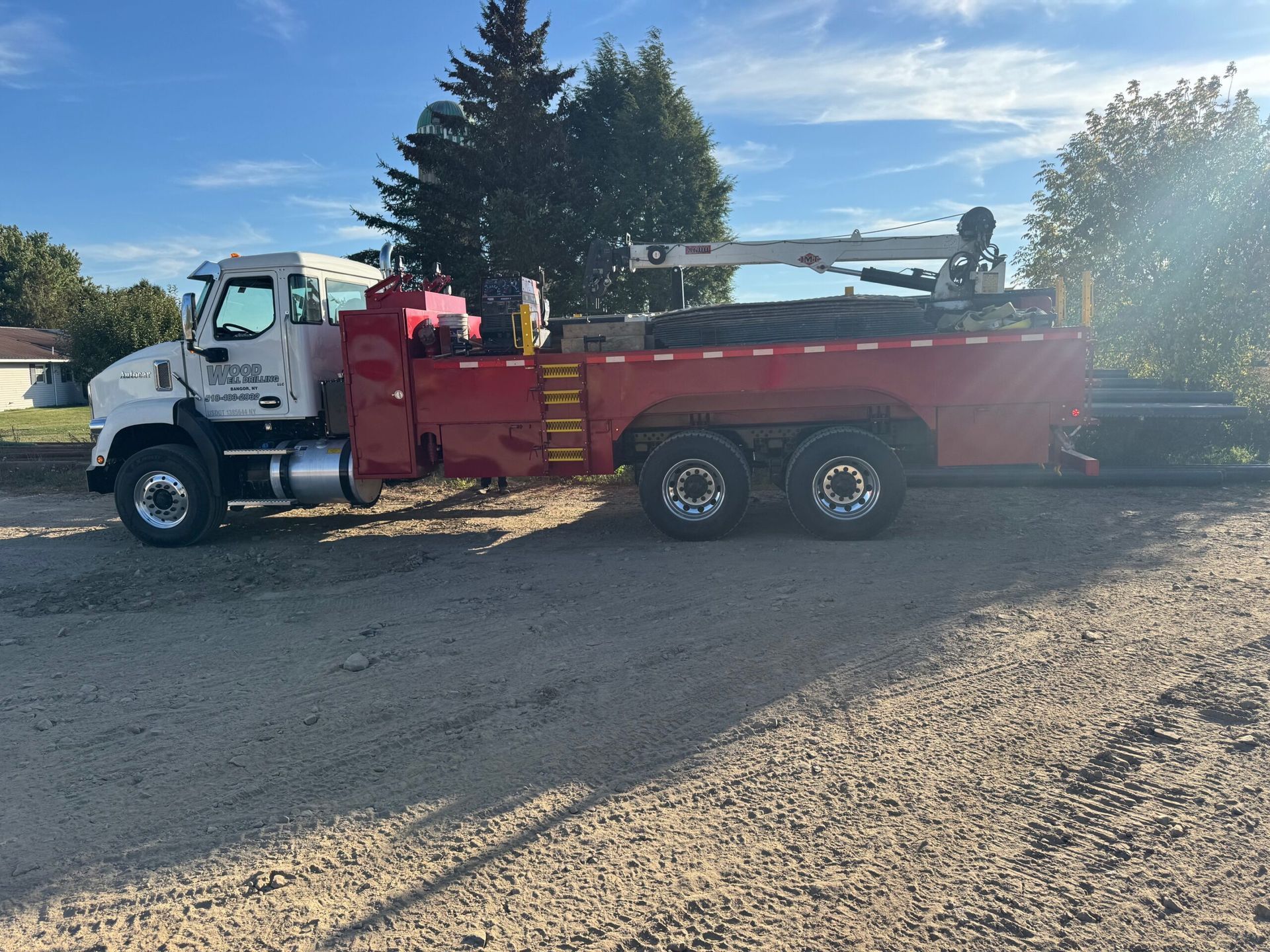 White utility truck with red dump bed parked on gravel in sunlight, near trees and a clear blue sky