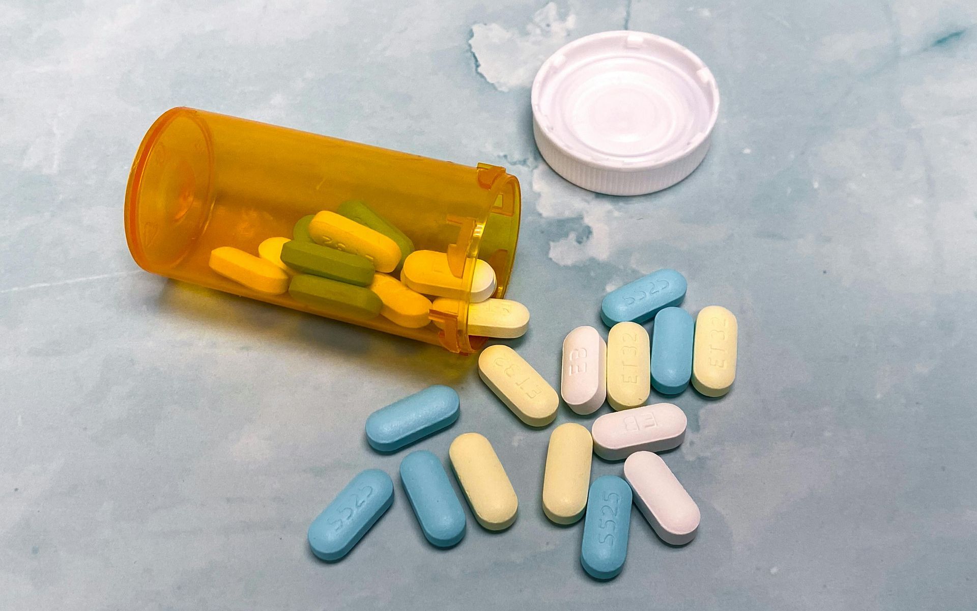 Orange pill bottle with pills spilled out: blue, yellow, and white pills on a light blue surface.