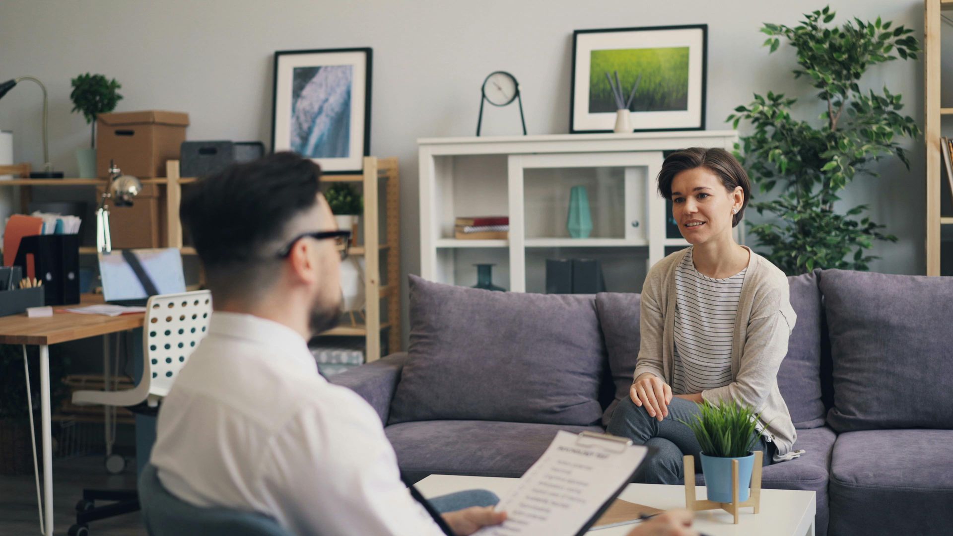 A therapist and patient in session. The patient sits on a couch, smiling. The therapist is holding a clipboard.