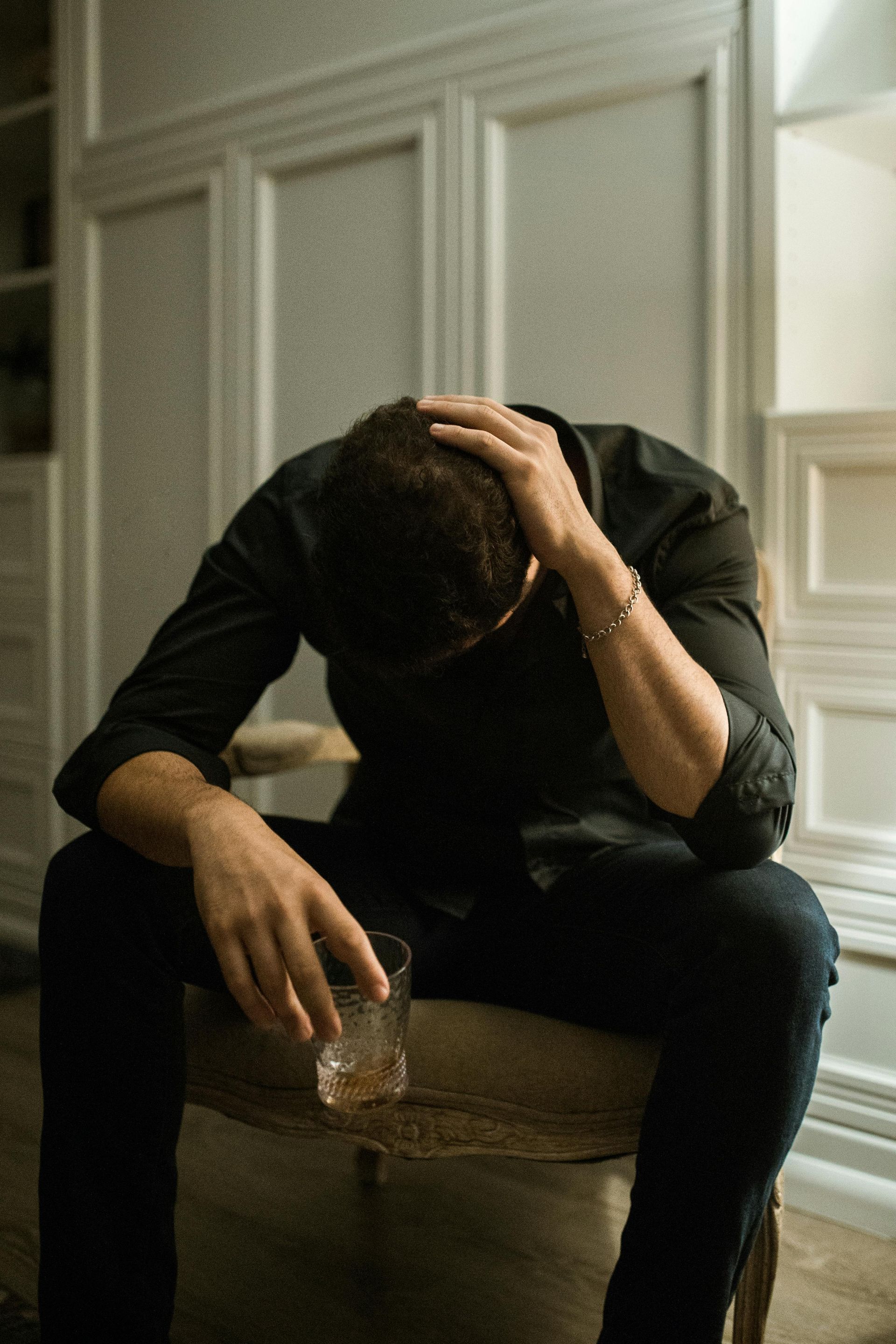 Man sitting with head in hands, holding a glass, looking distraught. Interior setting with white cabinetry.