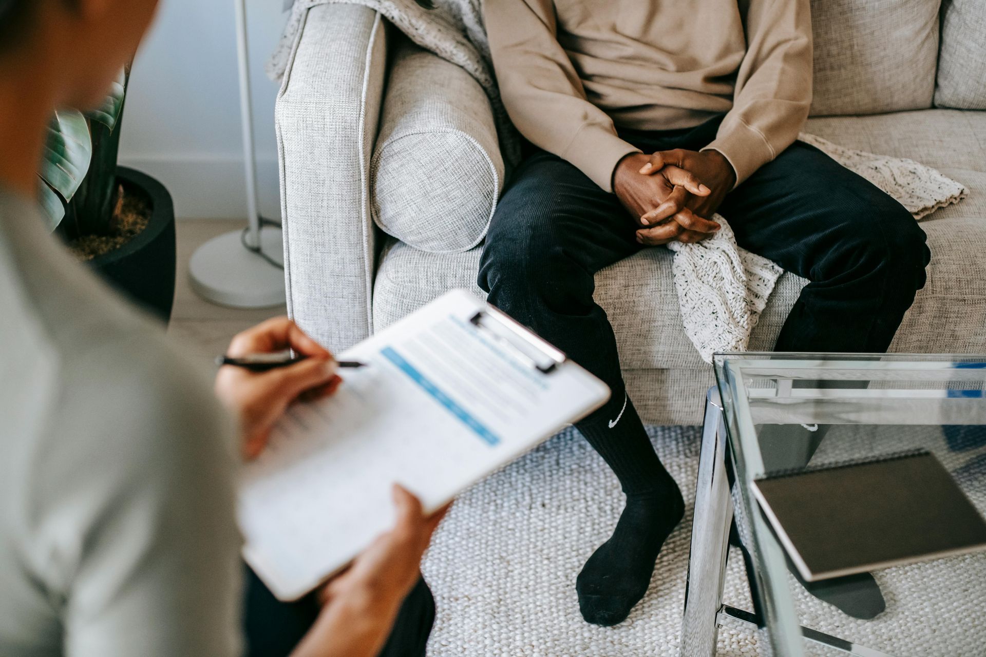 Person with a clipboard interviewing another person seated on a couch; indoors.