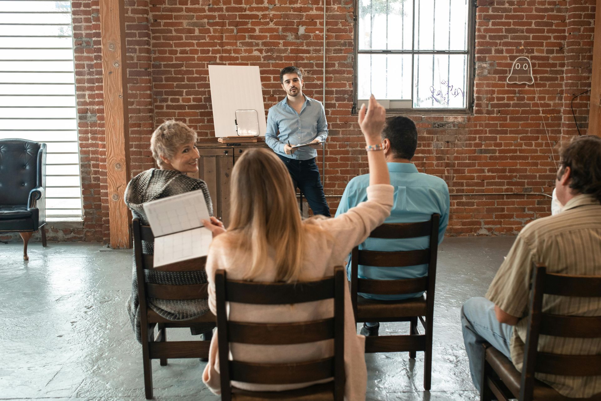 Man presents to group; woman raises hand to ask a question in a loft-style space.