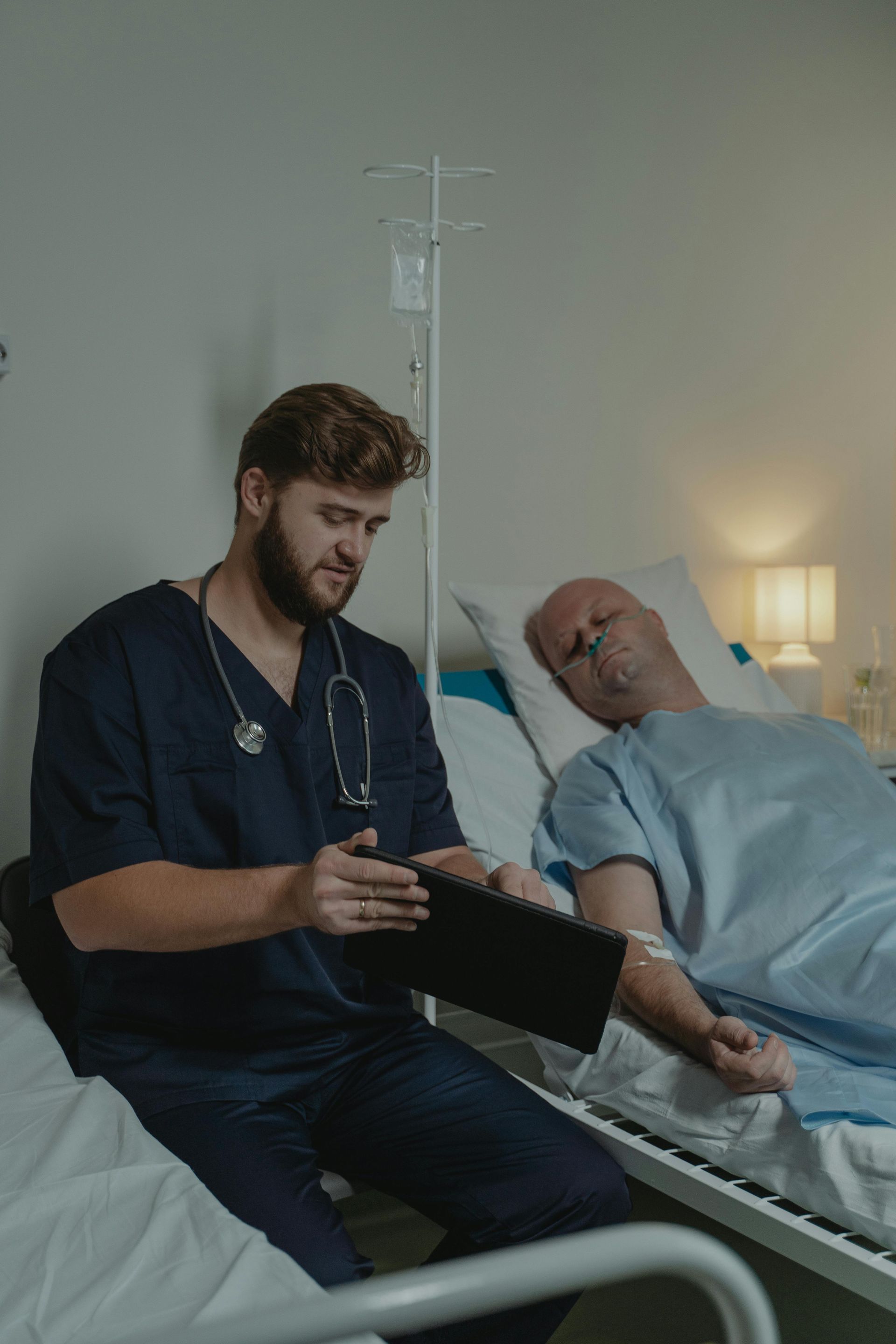 Nurse in scrubs reviews tablet, next to patient in hospital bed with IV.