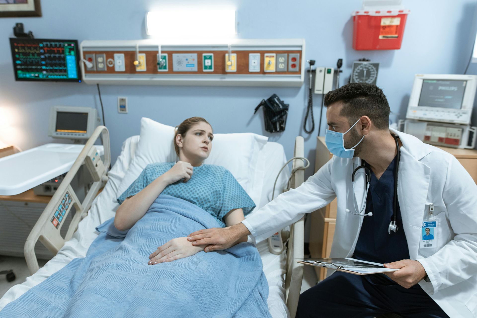 Doctor in mask examines patient in hospital bed; monitor shows vitals.