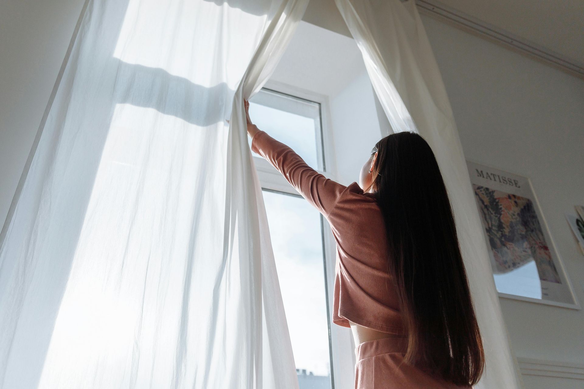 Woman opening sheer white curtains, revealing a bright window; interior shot.