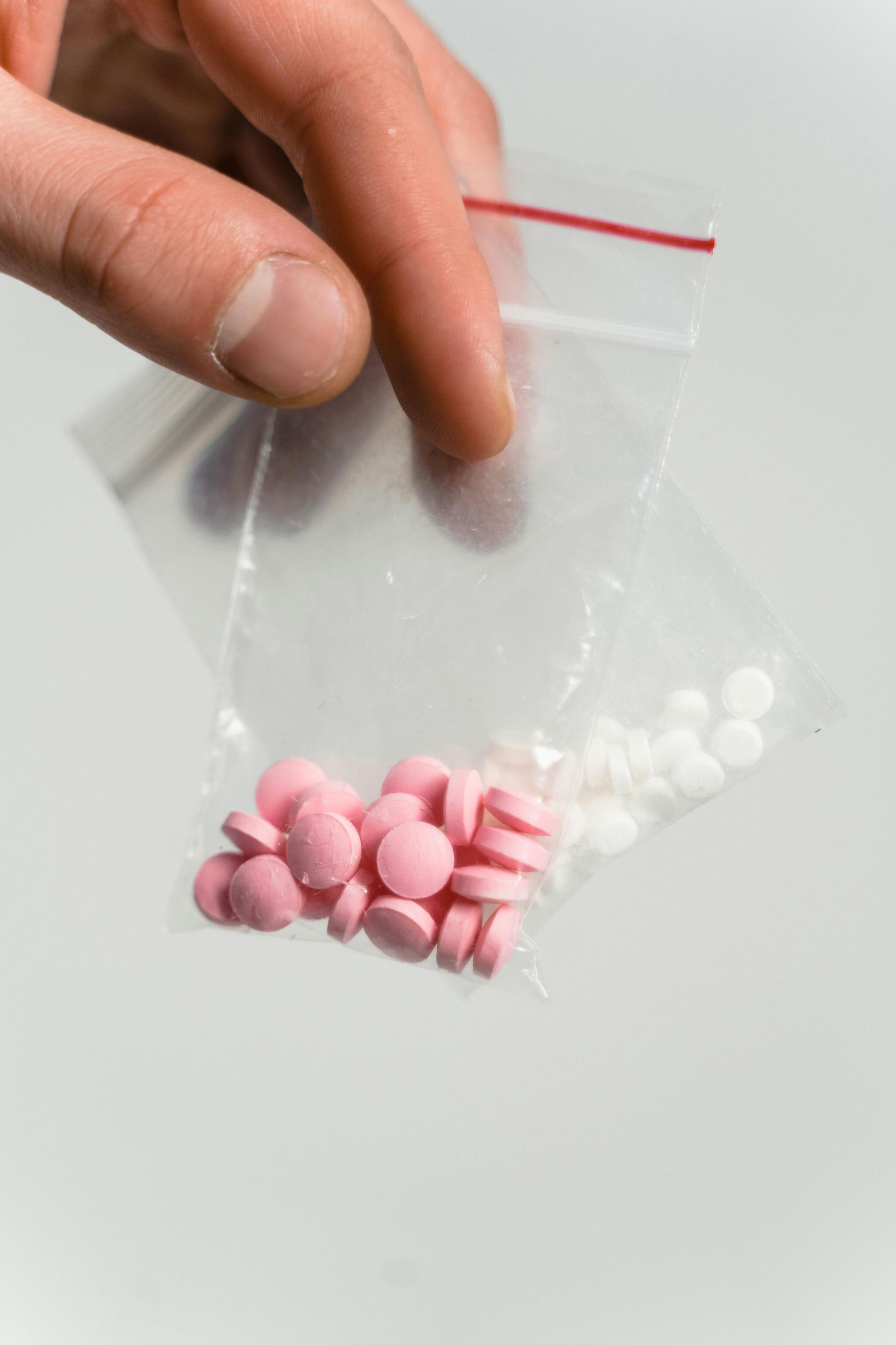 Hand holding a clear plastic bag containing pink and white pills on a light background.