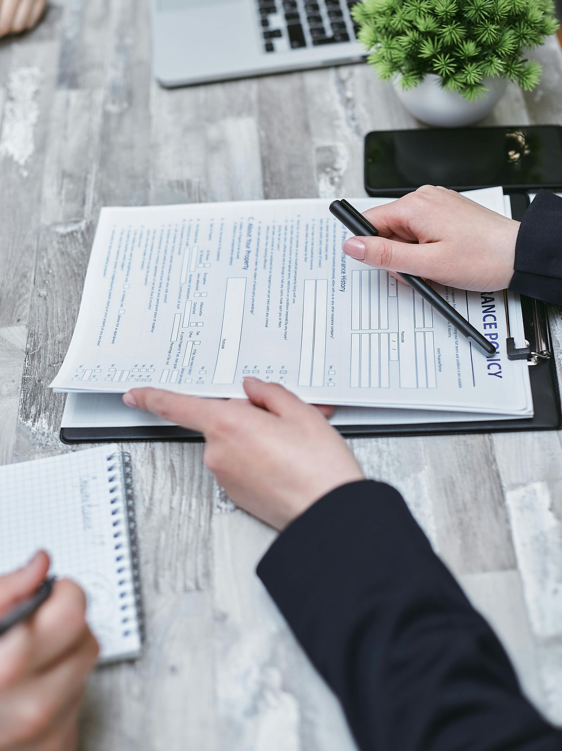 Hands pointing at a document on a desk, next to a notebook, pen, and a laptop.