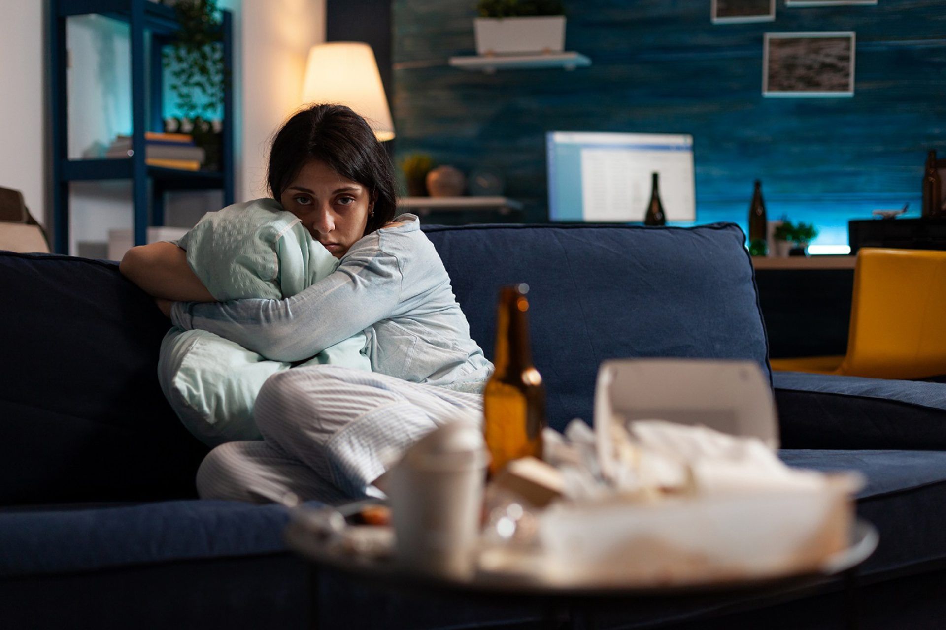 Person on a couch, holding pillow, looking sad. Table in foreground with alcohol and tissues. Dark, indoor setting.