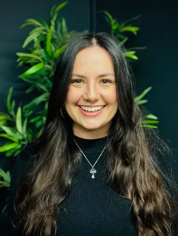 Woman with long dark hair, smiling broadly, wearing a black sweater and a necklace, in front of greenery.