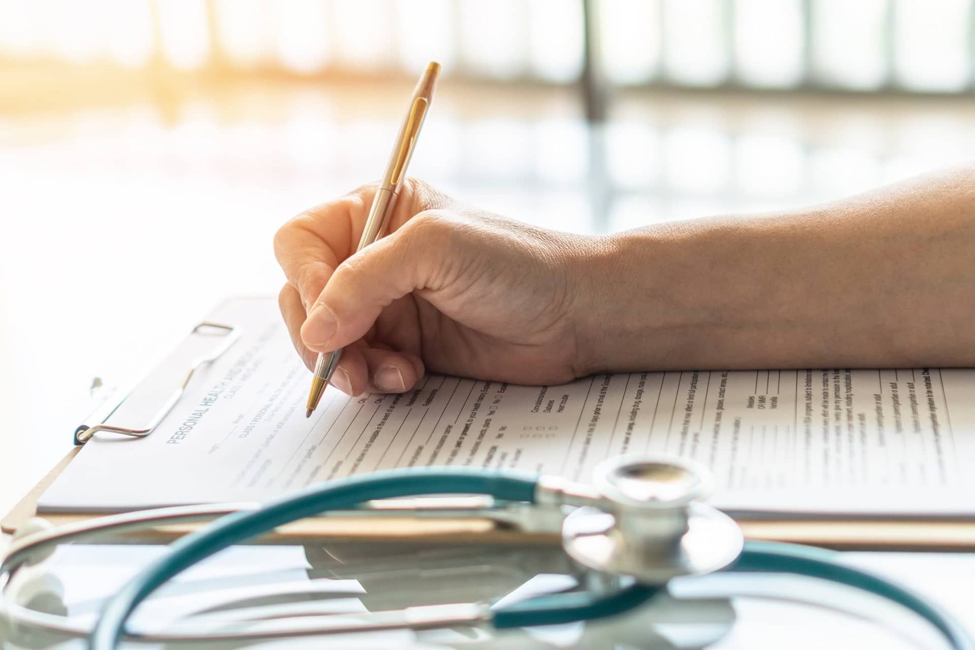 Hand writing on document on clipboard, stethoscope in foreground.