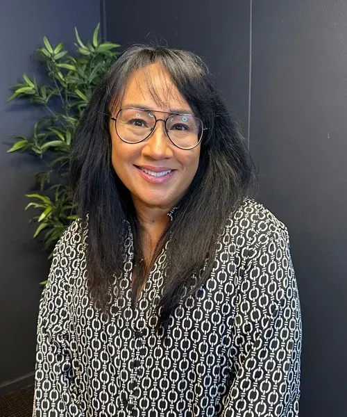 Woman with glasses smiles at the camera, wearing a patterned blouse. Dark background with a green plant.