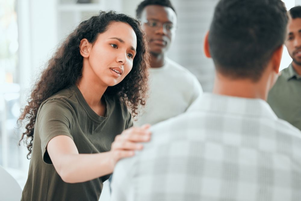 Woman comforts a person, hand on their shoulder, in a support group. Other people watch.