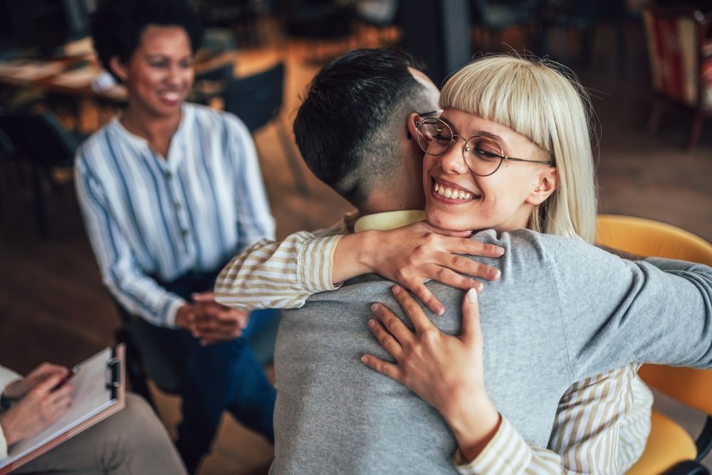 Two people hugging, smiling; person in background watches.