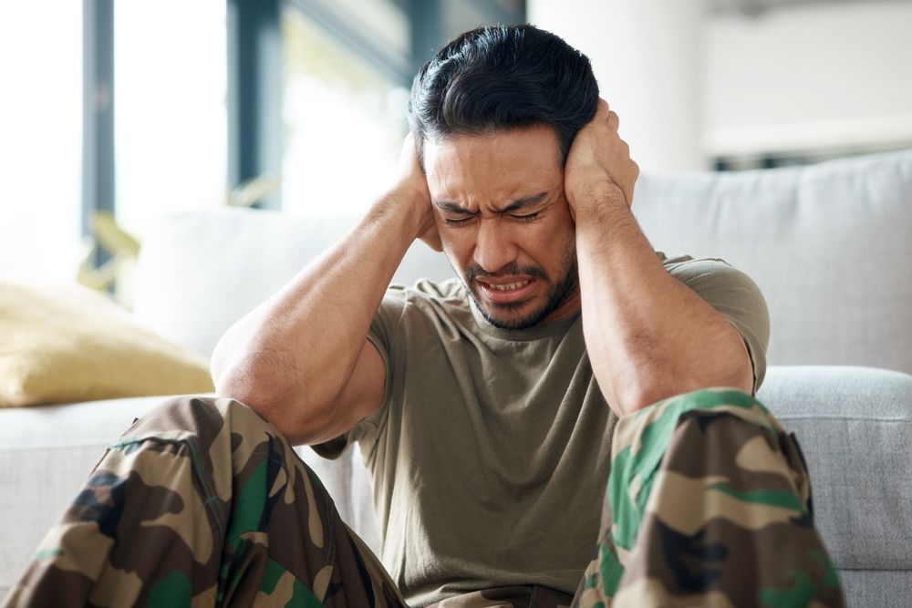 Man in camouflage pants clutches head in pain, sitting near a couch.