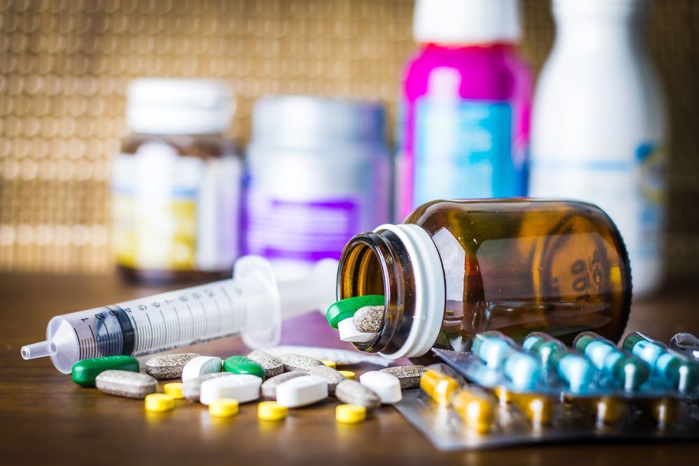 Pills and capsules spilled from a brown bottle; syringe, blister packs, and medicine bottles on table.