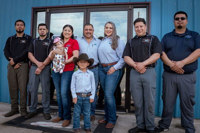 Seven people stand outside a blue building, posing together, with a child in a cowboy hat in front.