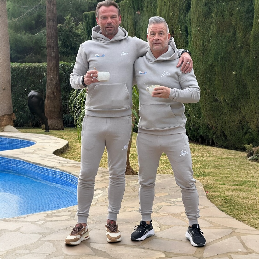Twee mannen in grijze trainingspakken staan bij een zwembad met bekers in hun handen.