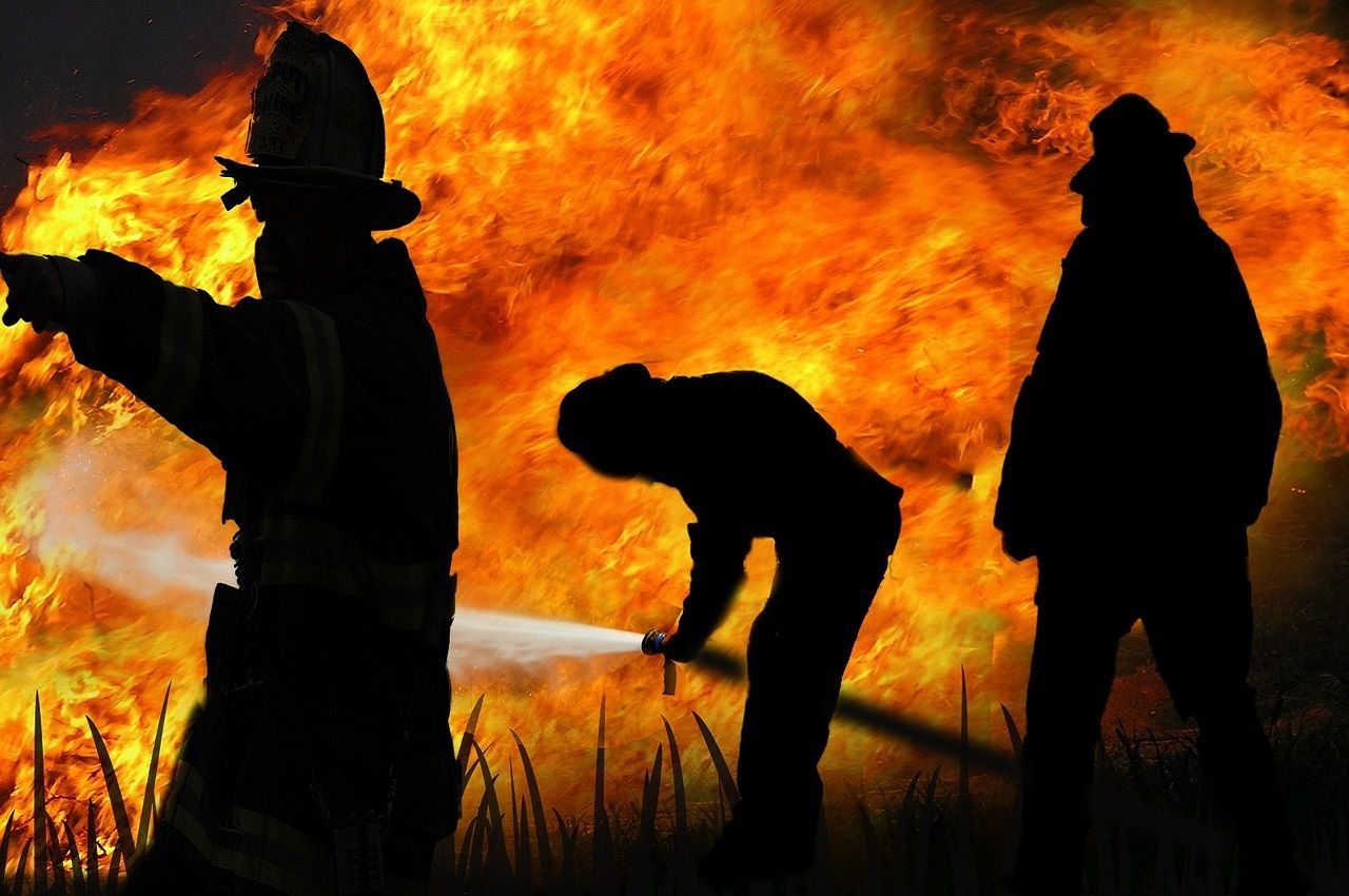 Brandweerlieden vormen silhouetten tegen een grote brand, terwijl ze water spuiten om de vlammen te blussen.