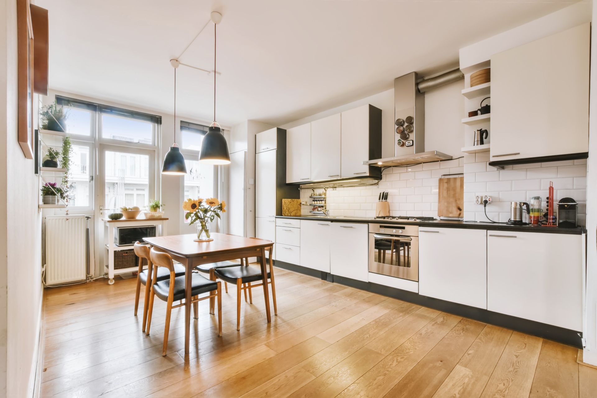 Modern kitchen with wooden floor, white cabinets, and a dining table with chairs. Sunlight streams in from the window.