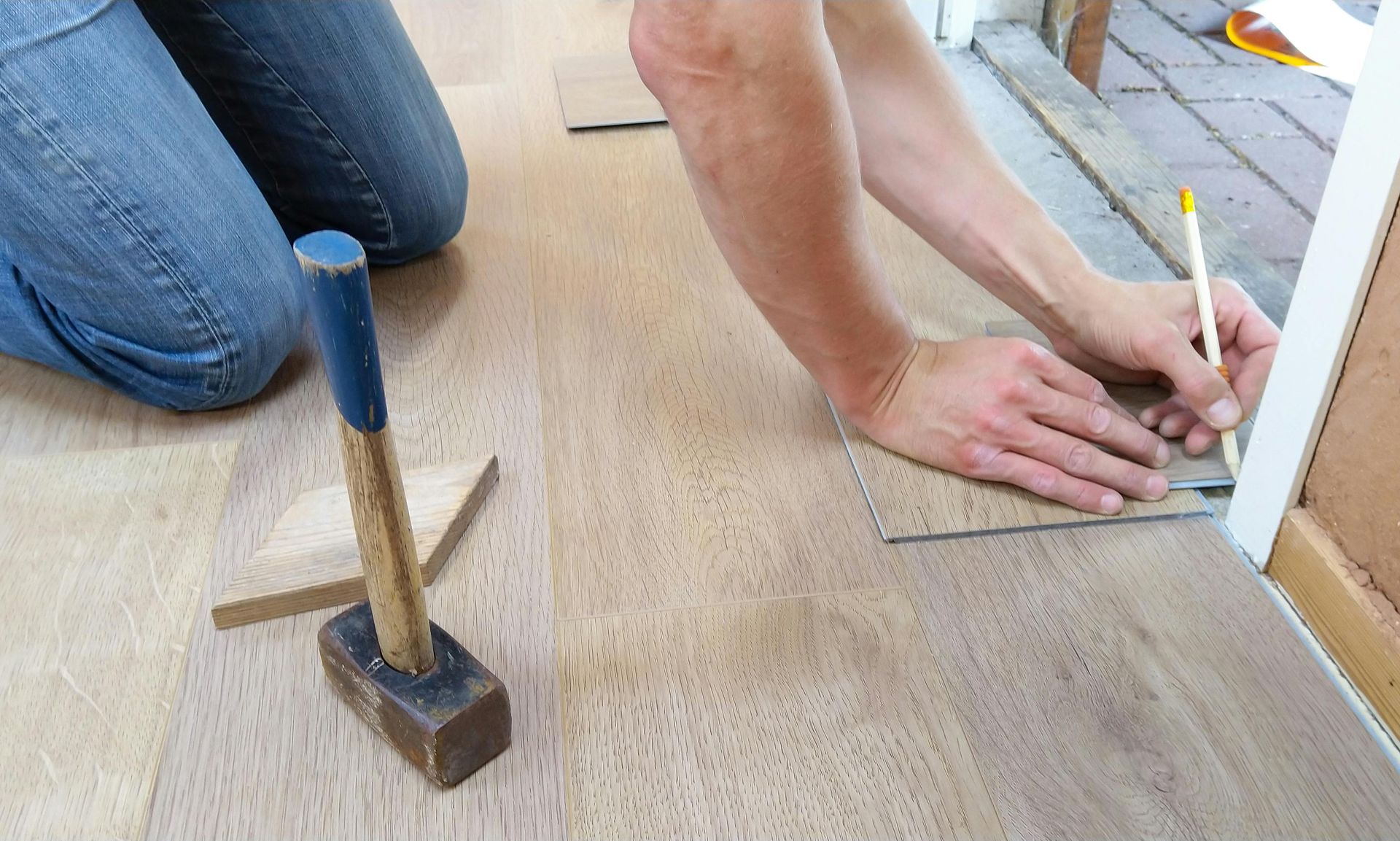 Person kneeling, marking flooring with a pencil and measuring tool, near a doorway. A hammer rests nearby.