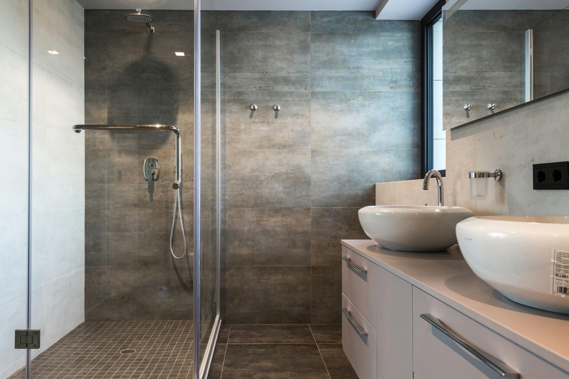 Modern bathroom with a glass-enclosed shower, two white sinks on a light-colored vanity, and grey tiled walls.