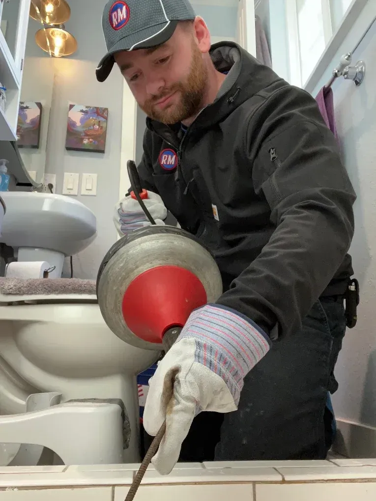 Plumber in bathroom unclogging drain with a cable, wearing a cap, gloves, and jacket.