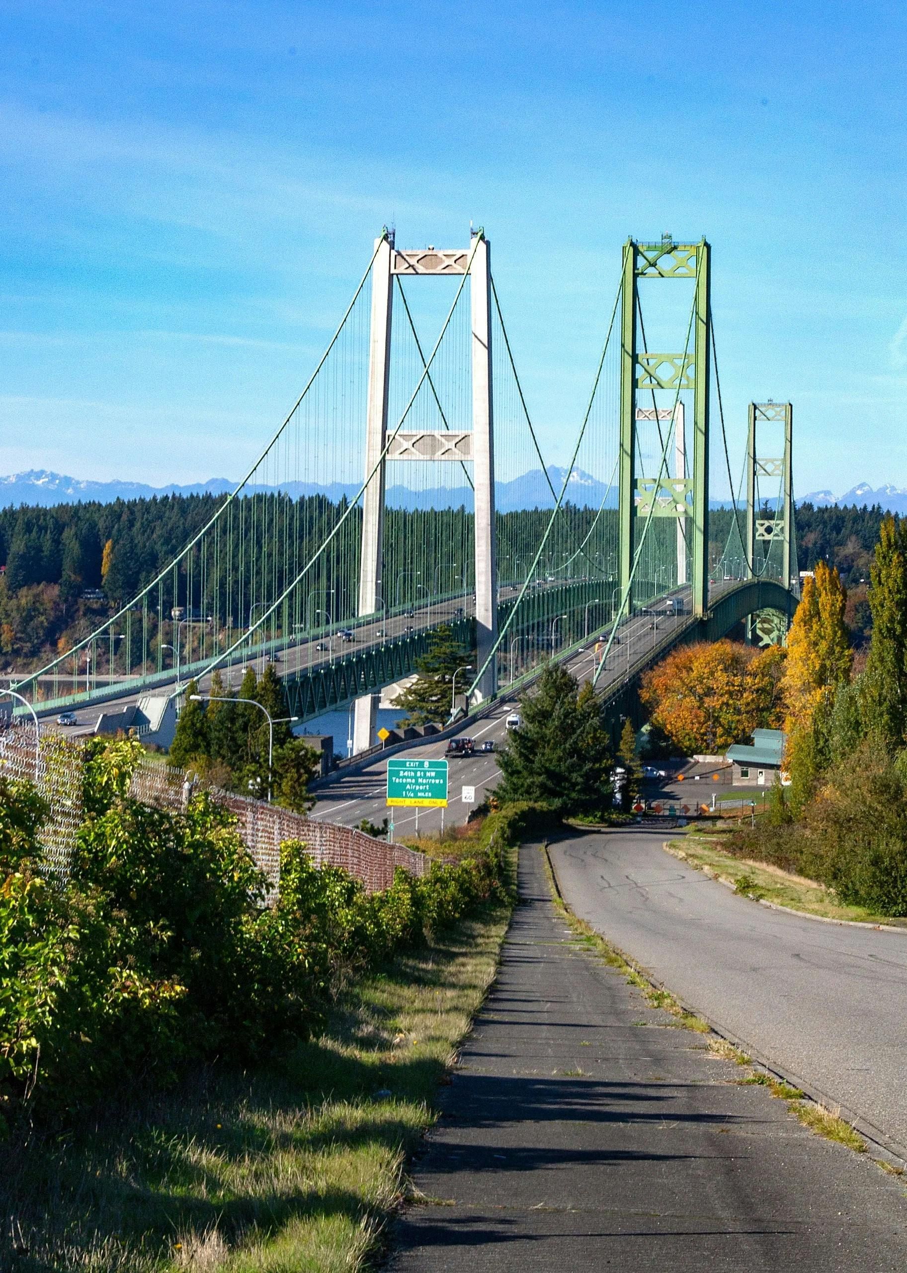 Tacoma Narrows Bridge, green and white suspension bridge spans water; roadway in foreground; sunny day.