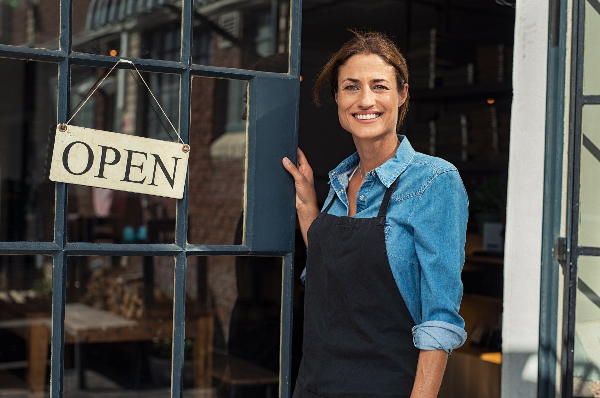 Woman wearing an apron, standing by her shop door, displayng an 'open' sign