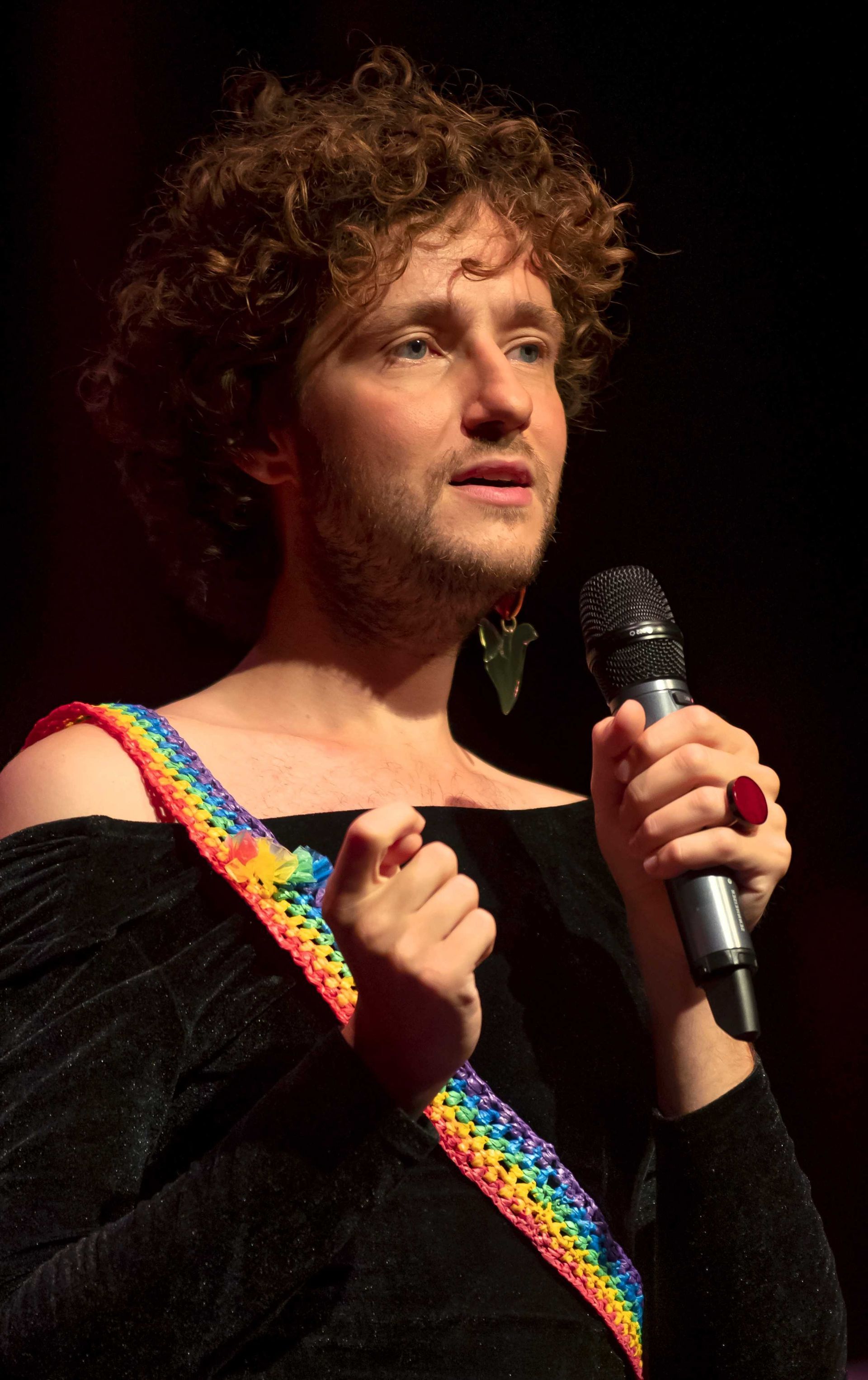 Rikkert van Huisstede, met krullend haar, houdt een microfoon vast en zingt in de Pride Show van Nederlied in Theater Het Zonnehuis in Amsterdam.