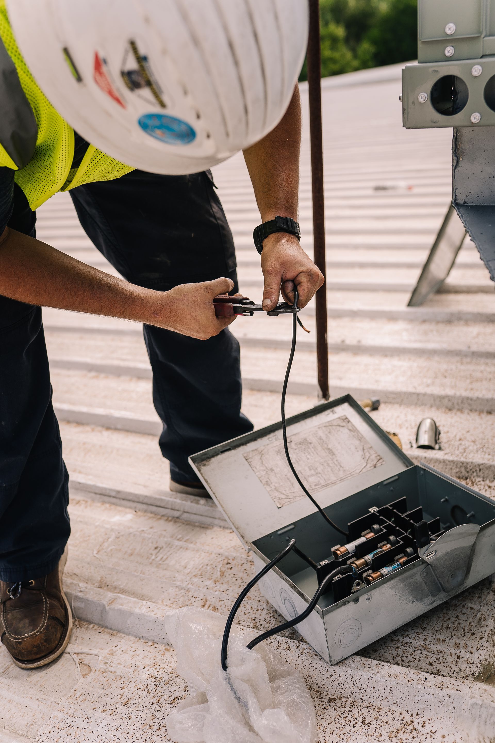 Construction worker on a rooftop, cutting wires in an electrical box, wearing a hard hat and safety vest.