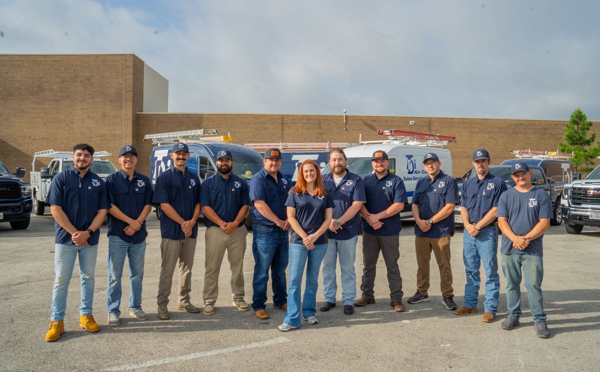 Group of HVAC technicians in blue shirts posing with vans in a parking lot.