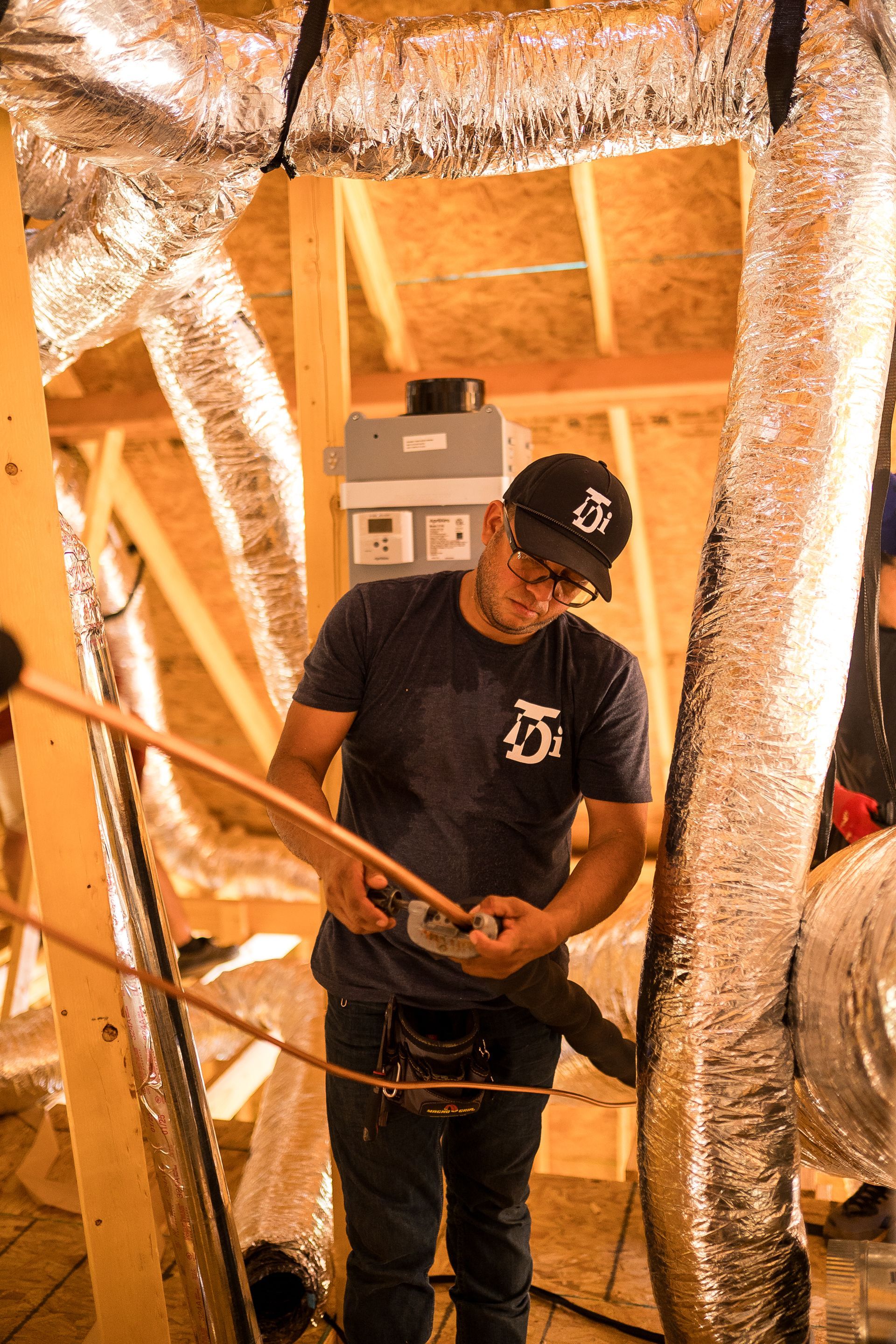 HVAC technician in an attic, working on copper pipes near ductwork and wood framing.