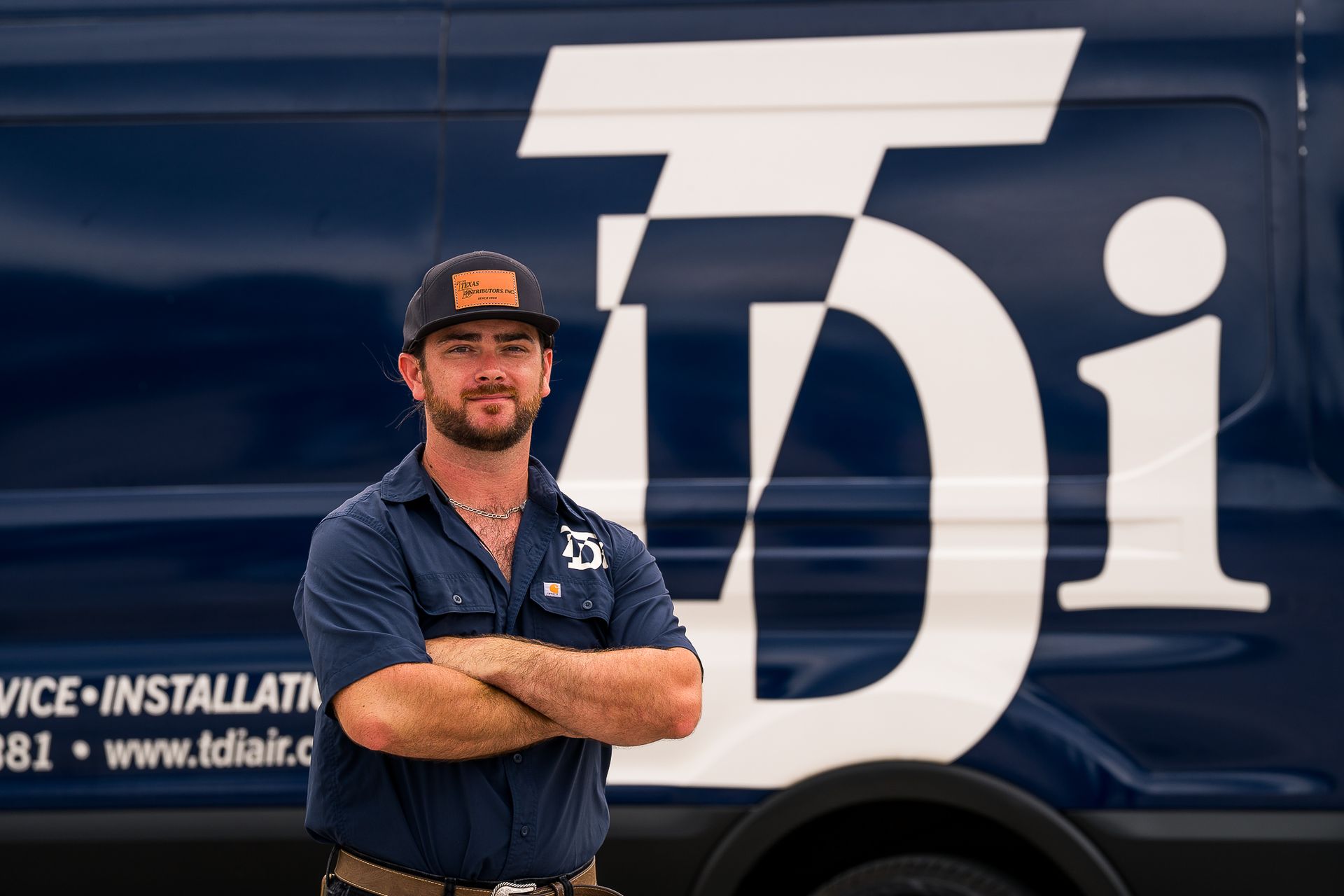 Man standing with arms crossed beside a dark blue service van with large white logo.